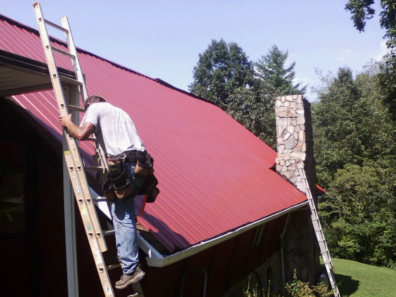 Roofer on a ladder beside a red metal roof and stone chimney — Soffit & Fascia Repair by Roofing Brothers in Wabasha